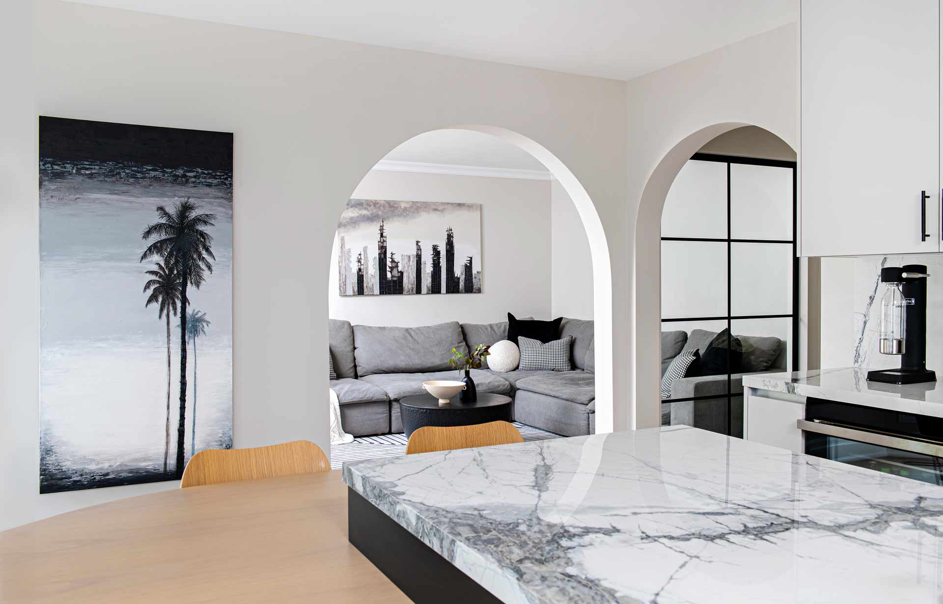 In the image by Unfold Creative Studio, the focal point is a luxurious kitchen with a seamless transition into the living room. The kitchen, adorned with high-end finishes, features a striking Black Palm painting by Alex Sauret on the left wall, adding a touch of artistic elegance to the space. Beyond the kitchen, framed by a beautiful arched entrance, lies the living room. The back wall of the living room is adorned with another captivating piece by Alex Sauret, titled "Disorder." This painting adds depth and intrigue to the living space, complementing the overall aesthetic and creating a harmonious visual flow between the kitchen and the living area. The two paintings are part of Alex Sauret's art portfolio and shop artwork.