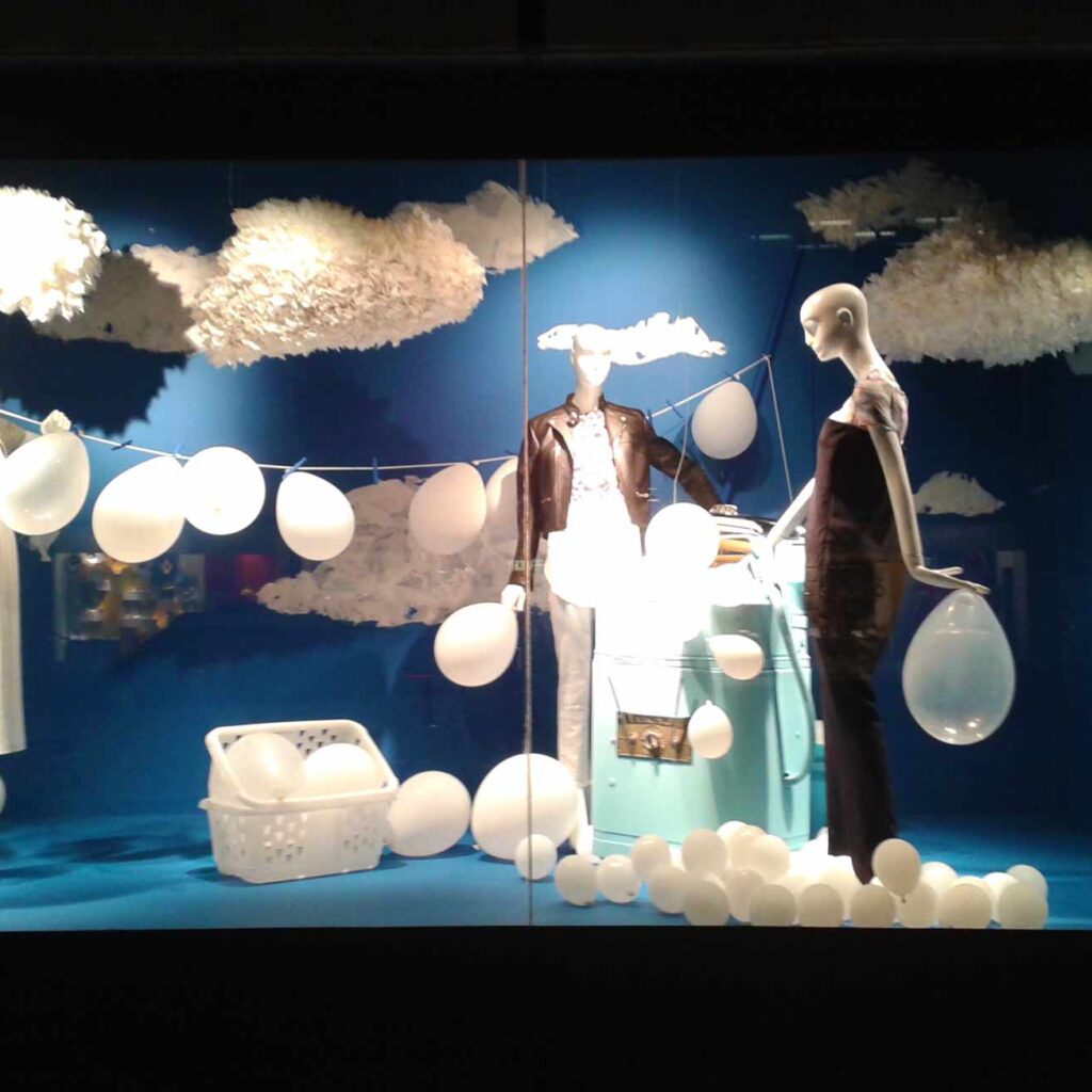 This image captures the Holt Renfrew Installation by Alex Sauret. The setting is imbued with blue hues, covering both the walls and the floor, creating a calm and dreamy atmosphere. Three mannequins, dressed in the brand's clothing, are the central focus, each holding white balloons that add an airy and whimsical element. Surrounding the mannequins are numerous floating, textured white clouds, enhancing the ethereal ambiance. A string stretches from one wall to another, adorned with a multitude of white balloons hanging from it. Smaller white balloons rest on the floor, scattered around and contributing to the playful feel of the installation. Additionally, white baskets filled with balloons are placed in the scene, further adding to the whimsical and light-hearted theme of the display.