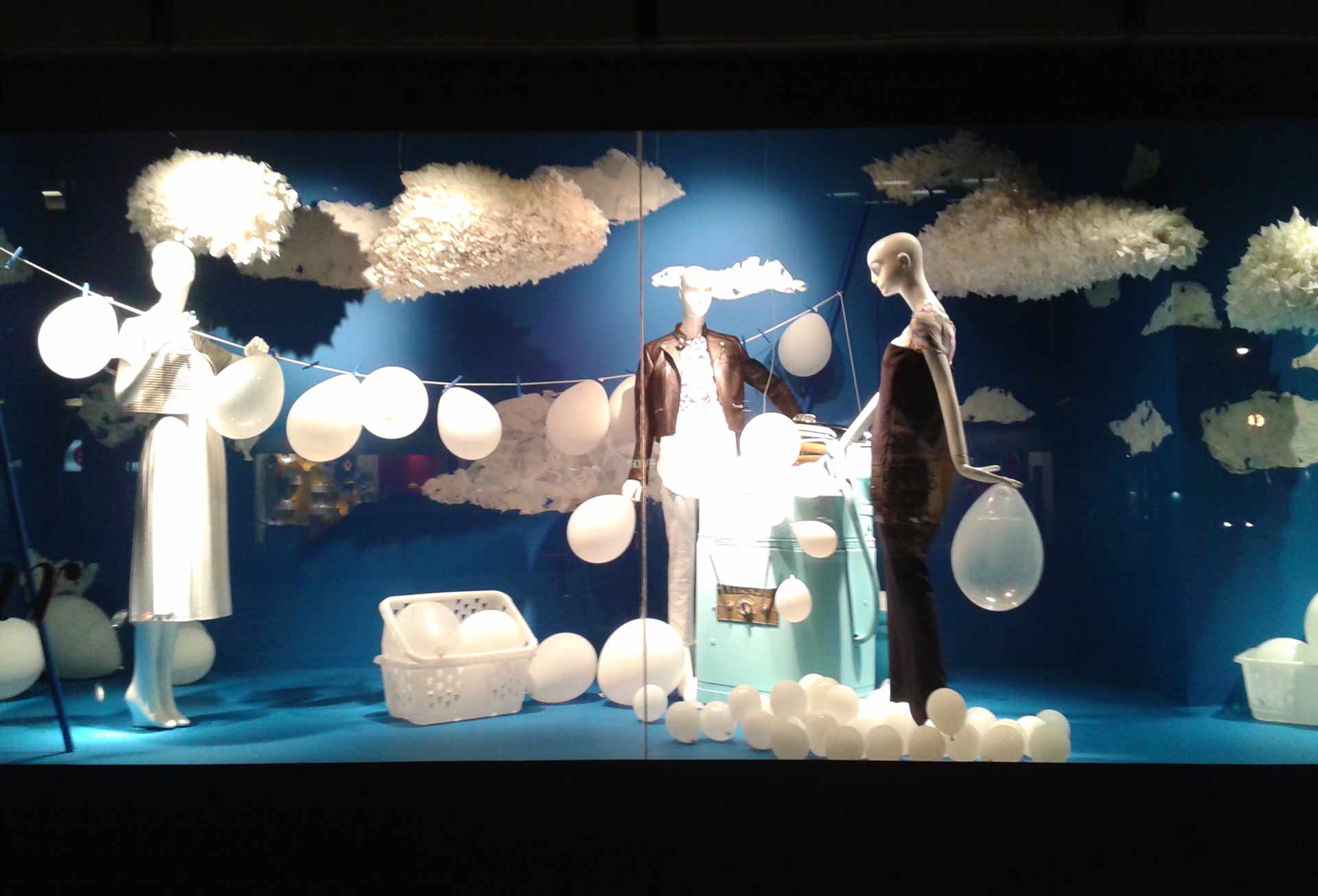 This image captures the Holt Renfrew Installation by Alex Sauret. The setting is imbued with blue hues, covering both the walls and the floor, creating a calm and dreamy atmosphere. Three mannequins, dressed in the brand's clothing, are the central focus, each holding white balloons that add an airy and whimsical element. Surrounding the mannequins are numerous floating, textured white clouds, enhancing the ethereal ambiance. A string stretches from one wall to another, adorned with a multitude of white balloons hanging from it. Smaller white balloons rest on the floor, scattered around and contributing to the playful feel of the installation. Additionally, white baskets filled with balloons are placed in the scene, further adding to the whimsical and light-hearted theme of the display.