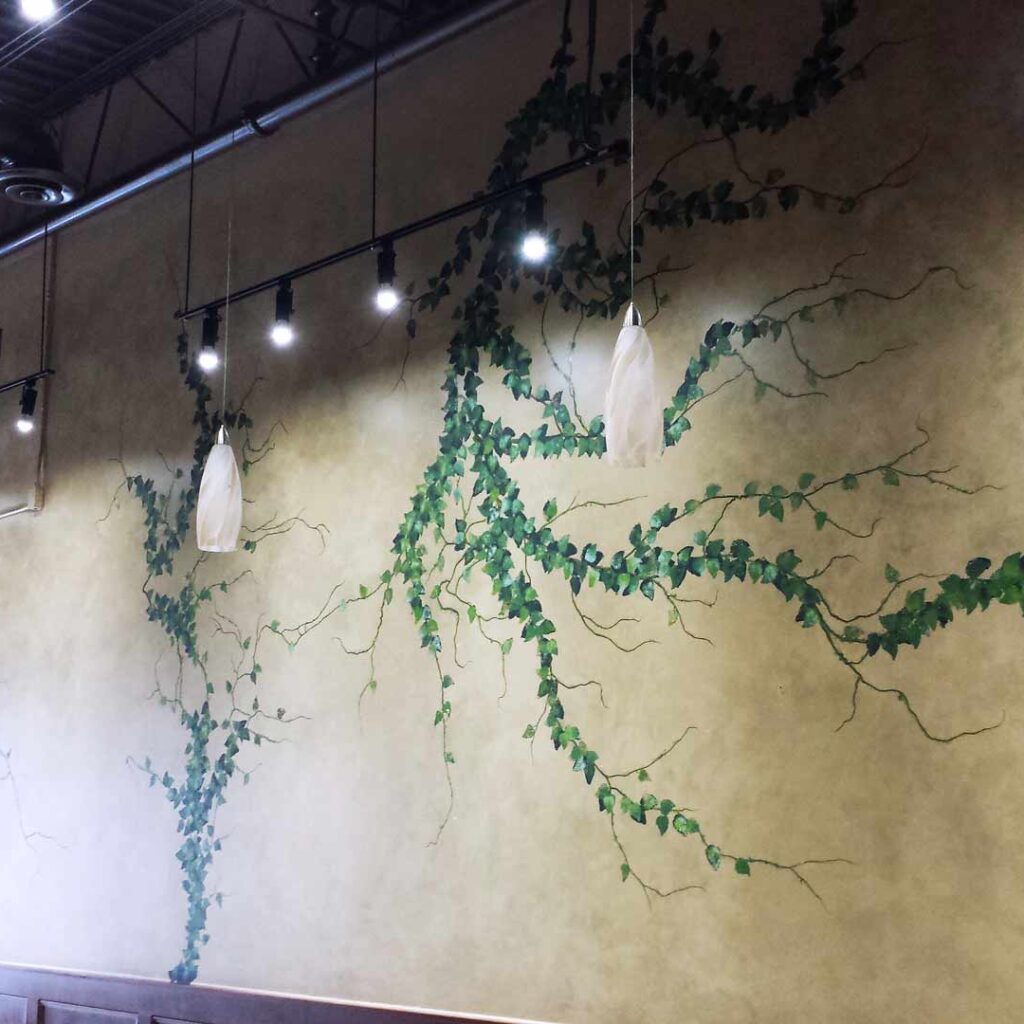 The image features a mural design by Alex Sauret created for Green Dragon CVR, a store. The mural's background is painted in gold hues, designed to mimic the look of marble. The 'marble' pattern includes three branches adorned with small green leaves, adding a natural element to the composition. The room has an industrial aesthetic, with a black ceiling where pipes and other infrastructure are visible. The mural is illuminated by hanging lights, which highlight its intricate details and vibrant colours, enhancing its visual impact within the store.