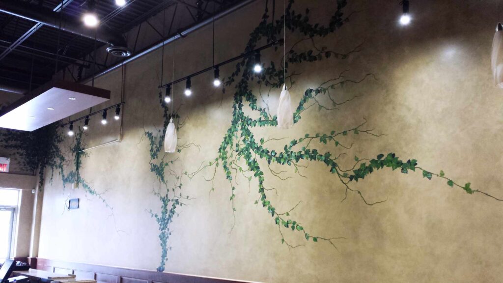 The image features a mural design by Alex Sauret created for Green Dragon CVR, a store. The mural's background is painted in gold hues, designed to mimic the look of marble. The 'marble' pattern includes three branches adorned with small green leaves, adding a natural element to the composition. The room has an industrial aesthetic, with a black ceiling where pipes and other infrastructure are visible. The mural is illuminated by hanging lights, which highlight its intricate details and vibrant colours, enhancing its visual impact within the store.